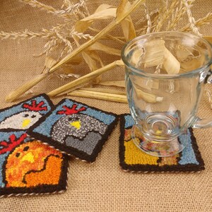May include: Three colorful crocheted coasters featuring a chicken design. The coasters are blue, gray, and orange. A clear glass mug is sitting on one of the coasters.