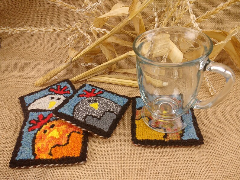 May include: Three colorful crocheted coasters featuring a chicken design. The coasters are blue, gray, and orange. A clear glass mug is sitting on one of the coasters.
