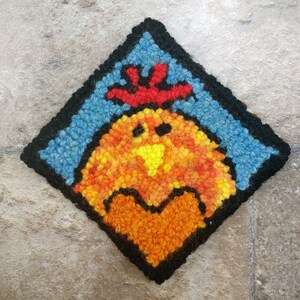 May include: A colorful needlepoint design of a rooster with a red comb and a heart in its chest. The rooster is surrounded by a blue background with a black border.