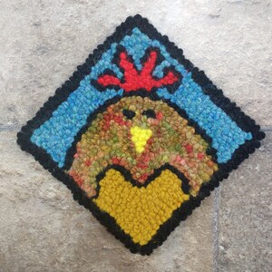 May include: A colorful wool rug depicting a rooster with a red comb and a yellow beak. The rooster is surrounded by a blue and black border.