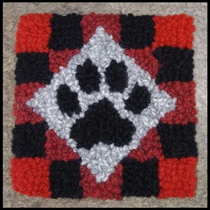 Pode incluir: Um tapete de l&atilde; vermelho, preto e branco com um desenho de pata no centro. O tapete &eacute; feito de l&atilde; e tem um padr&atilde;o xadrez.