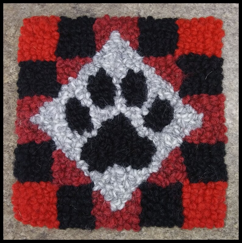 Pode incluir: Um tapete de l&atilde; vermelho, preto e branco com um desenho de pata no centro. O tapete &eacute; feito de l&atilde; e tem um padr&atilde;o xadrez.