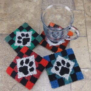 Pode incluir: Quatro  apoios de copo de croch&ecirc; feitos &agrave; m&atilde;o com um design de pegada de pata no centro. Os apoios de copo s&atilde;o em padr&otilde;es xadrez vermelho, verde, azul e laranja. Uma caneca de vidro transparente est&aacute; em cima de um dos apoios de copo.