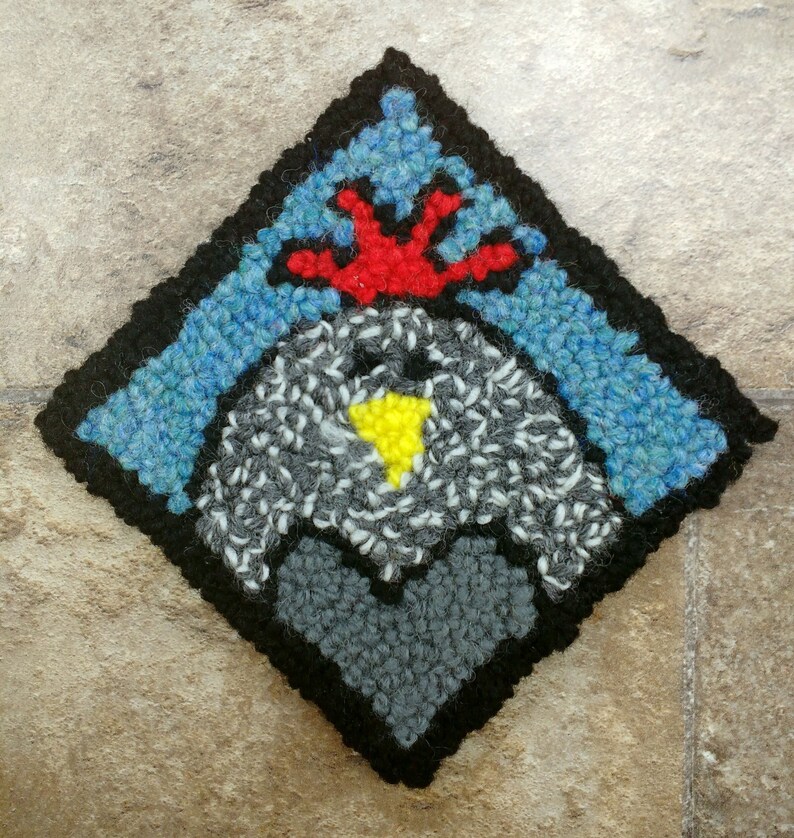 May include: A diamond-shaped rug with a black border and a gray, white, and yellow heart in the center. The heart is surrounded by a blue and red design.