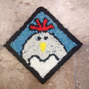 May include: A white, black, and red needlepoint rug with a stylized image of a chicken's head. The chicken has a red comb, a yellow beak, and black eyes.