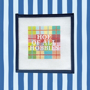 Peut inclure: Une toile de canevas carr&eacute;e pr&eacute;sentant un motif &eacute;cossais color&eacute; dans des tons de jaune, bleu, vert et rouge. Le texte "HOE OF ALL HOBBIES" est brod&eacute; en blanc au centre du motif. La toile est entour&eacute;e d'une bordure bleu marine fonc&eacute;.