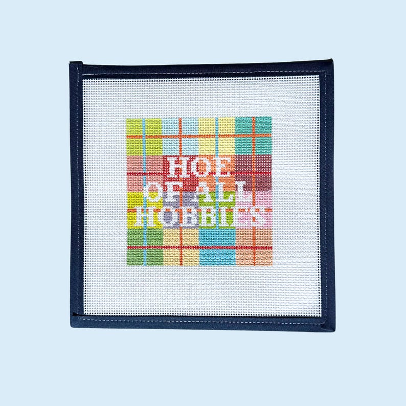 Peut inclure: Une toile de canevas carr&eacute;e avec un fond &agrave; grille blanche et un motif &eacute;cossais multicolore au centre. Le texte "HOE OF ALL HOBBIES" est brod&eacute; en blanc sur le motif &eacute;cossais. La toile pr&eacute;sente une bordure en tissu bleu marine fonc&eacute;.