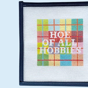 Peut inclure: Une toile de canevas carr&eacute;e pr&eacute;sentant un fond &agrave; carreaux color&eacute;s dans des tons de vert, jaune, bleu et rouge. Le texte blanc "HOE OF ALL HOBBIES" est brod&eacute; au centre. La toile poss&egrave;de une bordure en tissu bleu marine fonc&eacute; sur le c&ocirc;t&eacute; gauche.