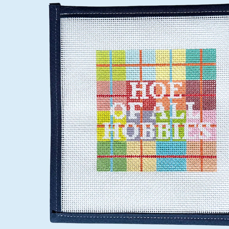 Peut inclure: Une toile de canevas carr&eacute;e pr&eacute;sentant un fond &agrave; carreaux color&eacute;s dans des tons de vert, jaune, bleu et rouge. Le texte blanc "HOE OF ALL HOBBIES" est brod&eacute; au centre. La toile poss&egrave;de une bordure en tissu bleu marine fonc&eacute; sur le c&ocirc;t&eacute; gauche.