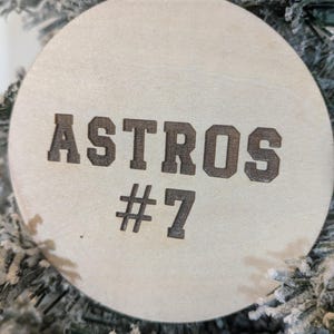 May include: A round, wooden Christmas ornament with the word "ASTROS" and the number "#7" etched in a dark brown color. The ornament is hanging on a frosted Christmas tree.