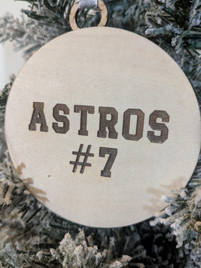May include: A round, wooden Christmas ornament with the word "ASTROS" and the number "#7" etched in a dark brown color. The ornament is hanging on a frosted Christmas tree.