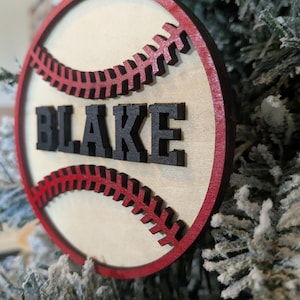 May include: A wooden baseball ornament with a red border and stitching. The name "BLAKE" is in black letters across the center. The ornament is hanging on a snowy Christmas tree.
