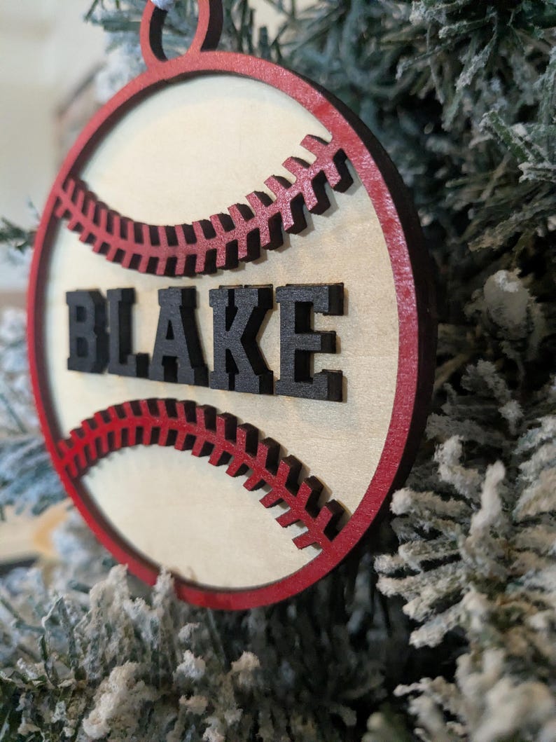 May include: A wooden baseball ornament with a red border and stitching. The name "BLAKE" is in black letters across the center. The ornament is hanging on a snowy Christmas tree.