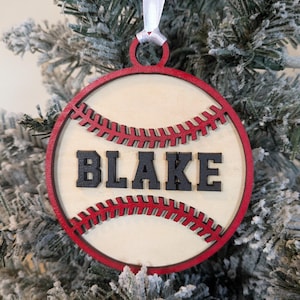 May include: A round, wooden baseball ornament with the name "BLAKE" in black letters. The ornament has a red border and red stitching details, and is hanging from a white ribbon. The ornament is displayed on a snowy Christmas tree.
