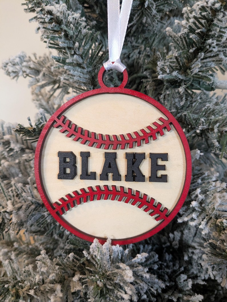 May include: A round, wooden baseball ornament with the name "BLAKE" in black letters. The ornament has a red border and red stitching details, and is hanging from a white ribbon. The ornament is displayed on a snowy Christmas tree.