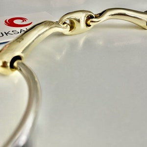 May include: A gold-coloured horse bit with a curved mouthpiece and a silver ring. The bit is designed for use in equestrian sports.