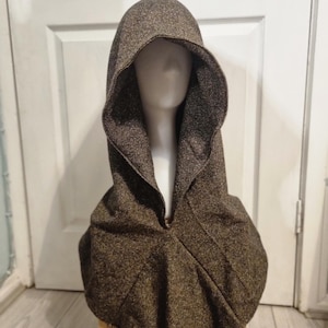 Könnte beinhalten: Ein brauner Kapuzenmantel aus strukturiertem Stoff. Der Umhang hat eine tiefe Kapuze und eine Öffnung vorne. Das Kleidungsstück wird auf einem Schaufensterkopf präsentiert, um seine Form und sein Design hervorzuheben. Der Umhang scheint ein warmes Oberbekleidungsstück zu sein.