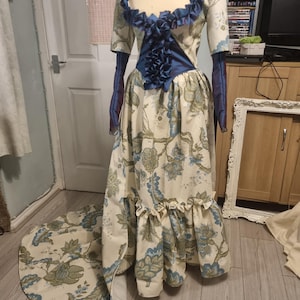 May include: A cream-colored gown with a floral pattern in blue and green, displayed on a mannequin. The dress features a ruffled neckline and bodice, with long, sheer blue sleeves. The skirt has a ruffled hem and a long train.