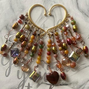 May include: A pair of gold-toned ear cuffs featuring cascading dangles of multi-colored beads in shades of amber, green, brown, and red. The jewelry includes faceted crystals, smooth round beads, and a prominent dark brown heart-shaped charm.