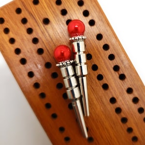 May include: Two red and silver game pieces with a pointed tip, sitting on a wooden game board with a grid of holes.