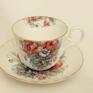 Puede incluir: Juego de taza y platillo blancos con diseño floral que incluye amapolas rojas, flores azules y blancas. La taza y el platillo tienen ribetes dorados. Un juego de té clásico.