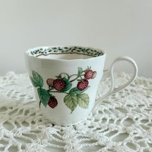 May include: White ceramic teacup with a curved handle. The cup is decorated with a raspberry and leaf design. The rim of the cup has a green and brown leaf pattern. The cup is sitting on a white lace doily.