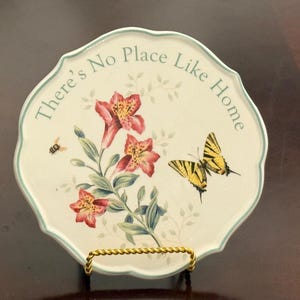 May include: Decorative ceramic plate with a scalloped edge, featuring a floral design with red and yellow flowers, a yellow butterfly, and a bee. The plate has the text "There's No Place Like Home" in a light blue font.