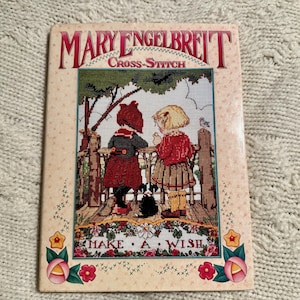 May include: A cross-stitch pattern book by Mary Engelbreit featuring two children in red and green clothing standing in front of a fence with a dog and the text "Make a Wish".