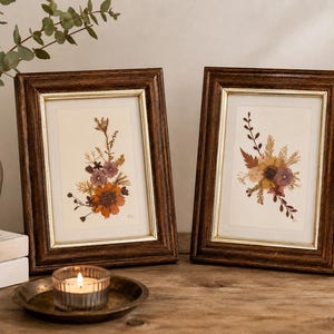 May include: Two rectangular picture frames with dark brown wood grain finish and thin gold inner borders. Each frame contains a cream-colored mat and a pressed flower arrangement featuring dried orange, purple, and brown botanical elements.