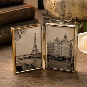 Puede incluir: Un marco de fotos doble de metal dorado con bisagras y dos paneles rectangulares. El panel izquierdo muestra una fotografía en blanco y negro de la Torre Eiffel en París, mientras que el panel derecho muestra una fotografía en blanco y negro de un edificio.