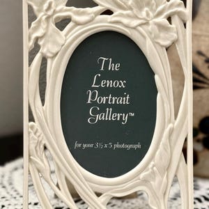 Puede incluir: Marco de fotos rectangular blanquecino con una abertura ovalada. El marco presenta un diseño floral y el texto "The Lenox Portrait Gallery" y "for your 3 1/2 x 5 photograph".