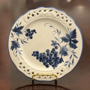 Puede incluir: Un plato de cerámica blanco con un borde azul y un diseño decorativo de uvas. El plato presenta un borde de celosía y está sostenido por un soporte dorado. Las uvas y las hojas están pintadas en tonos azules.