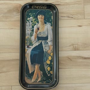 May include: Vintage Coca-Cola serving tray with a black border. The tray features a woman in a dress holding a glass of Coca-Cola. The Coca-Cola logo is at the top of the tray.