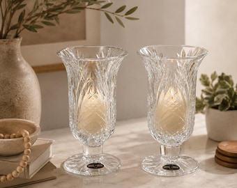 May include: A pair of clear crystal hurricane candle holders featuring a flared rim, intricate vertical cut patterns, and a sturdy circular base. Each piece contains a cream-colored pillar candle and displays a small black label on the base.