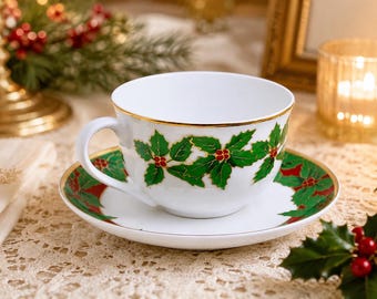 May include: A white porcelain teacup and saucer set featuring a festive holly pattern with vibrant green leaves and small red berries. The rim of the cup and the edge of the saucer are finished with a thin, elegant gold trim, adding a refined detail.