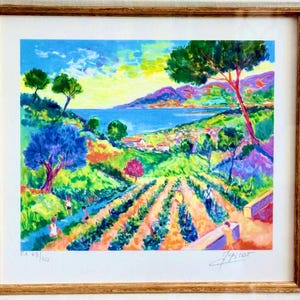 May include: A framed landscape painting with a gold frame. The artwork depicts a colorful coastal scene with a blue sea, green hills, and a vineyard. The sky is a mix of yellow and blue. The painting is signed and numbered.