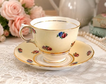 May include: A cream-colored porcelain teacup and saucer set featuring a delicate floral pattern of small red, blue, and purple roses. The rim of the cup and the edge of the saucer are accented with a thin, polished gold trim, adding a refined finish.
