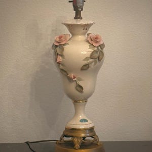 May include: A cream-colored ceramic table lamp base with a bulb socket at the top. The body features raised, three-dimensional pink rose blossoms and green leaves. The lamp stands on a circular, ornate gold-toned metal base with decorative feet.