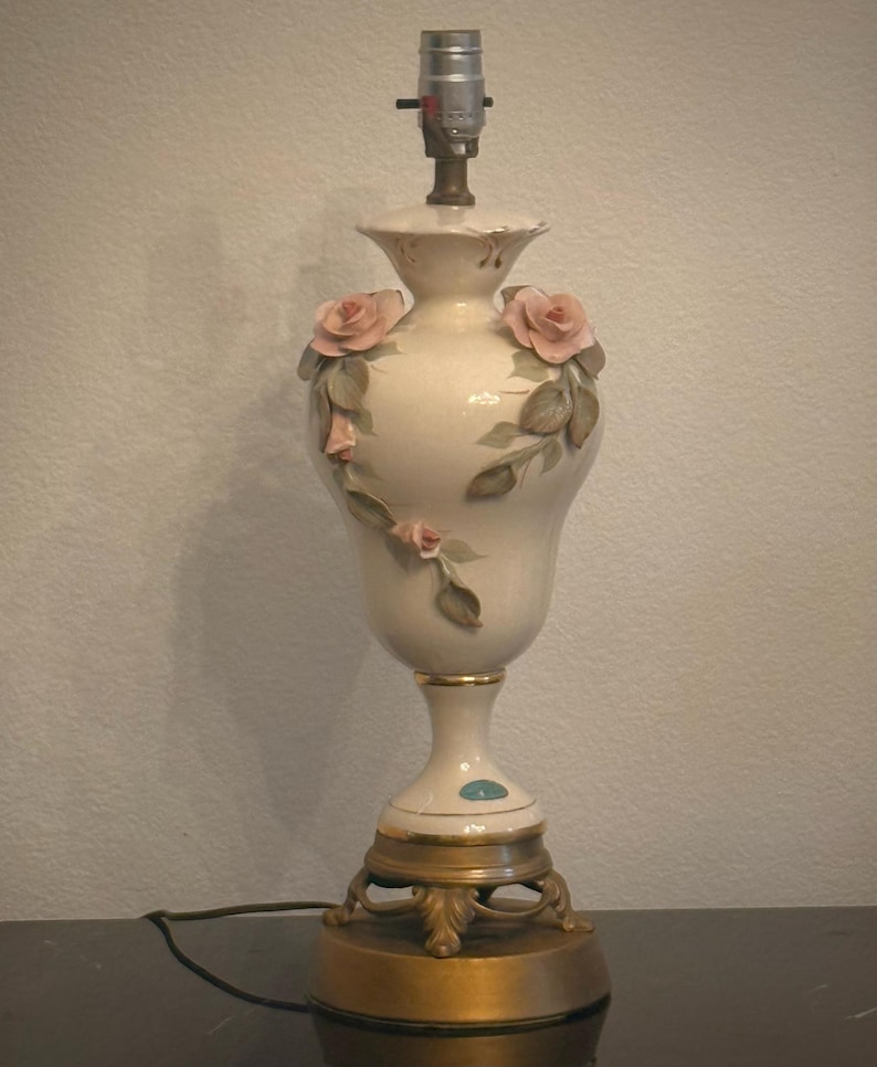 May include: A cream-colored ceramic table lamp base with a bulb socket at the top. The body features raised, three-dimensional pink rose blossoms and green leaves. The lamp stands on a circular, ornate gold-toned metal base with decorative feet.