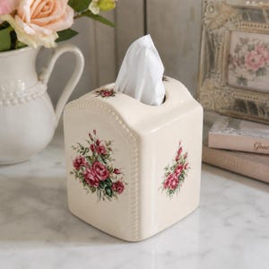 May include: A cream-colored ceramic tissue box cover with a rectangular shape and rounded top edges. The exterior features a delicate beaded border detail and a floral pattern of pink roses with green leaves on the front and side panels.