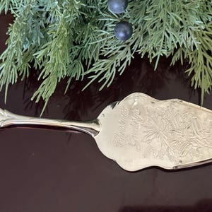 Op de afbeelding: Zilveren taartschep met een sierlijk handvat en een decoratief blad. Het blad heeft een gegraveerd bloemmotief en de woorden "Christmas Greetings". Groen en bessen staan op de achtergrond.