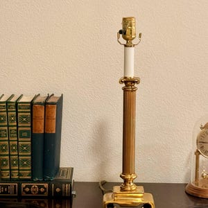May include: A gold-colored table lamp with a fluted column design and a square base. The lamp has a white central section and a brass-colored lamp holder. The lamp is on a dark surface, with books and a clock in the background.