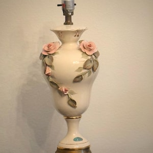 May include: A cream-colored ceramic table lamp base with a bulb socket at the top. The body features three-dimensional pink rose blossoms and green leaves applied to the surface. The base is supported by a gold-toned metal stand with ornate feet.