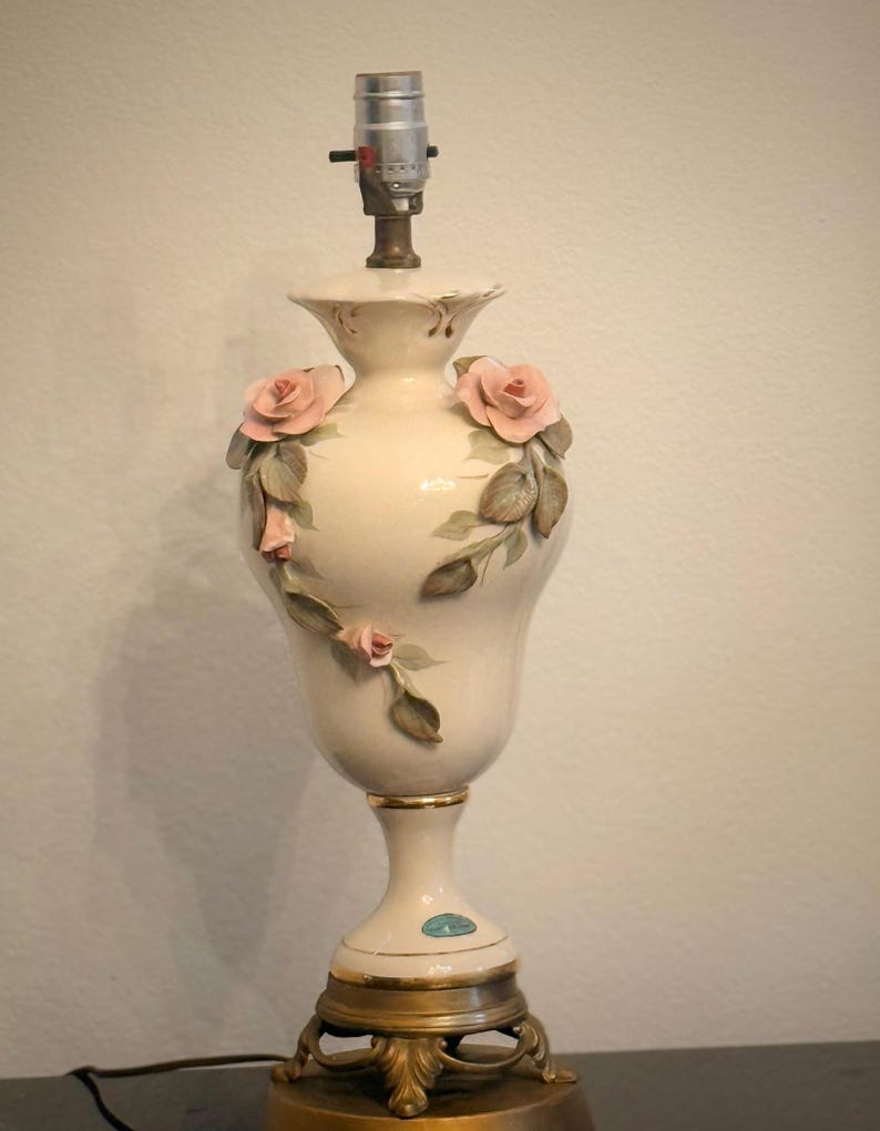 May include: A cream-colored ceramic table lamp base with a bulb socket at the top. The body features three-dimensional pink rose blossoms and green leaves applied to the surface. The base is supported by a gold-toned metal stand with ornate feet.
