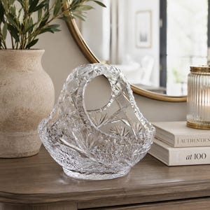 Op de afbeelding: Een heldere, kleurloze glazen mand met een ingewikkeld, veelzijdig geslepen kristalpatroon met ster- en diamantmotieven. De mand heeft een gebogen, in het ontwerp geïntegreerd handvat en een ronde voet die het licht door het oppervlak weerkaatst.