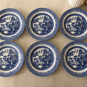 Pode incluir: Um conjunto de seis pratos redondos de cerâmica com um padrão de salgueiro em azul e branco. Cada prato apresenta uma ilustração detalhada de paisagem com pagodes, árvores e pássaros, rodeada por uma borda geométrica decorativa no rebordo.