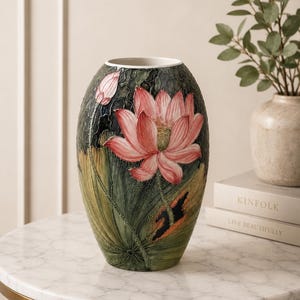 Könnte beinhalten: Eine ovale Keramikvase mit einem Reliefdesign aus einer großen rosa Lotusblüte und einer kleineren Knospe. Die Oberfläche ist mit strukturierten grünen Blättern und dunklen, wirbelnden Mustern verziert. Die Vase hat einen weißen Innenrand.