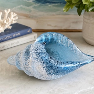 May include: A decorative glass conch shell bowl featuring a gradient of deep blue to light blue with a textured, frosted, and speckled finish. The shell has a smooth, glossy interior and a wide, open aperture, showcasing a coastal aesthetic.