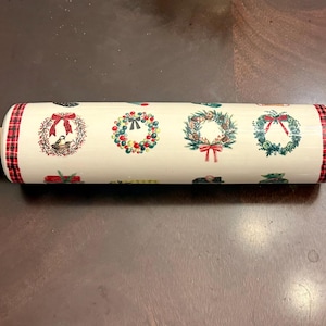 May include: A wooden rolling pin with a cream-colored barrel decorated with Christmas wreaths and red plaid accents. The rolling pin has wooden handles on each end. The wreaths are green, red, and white.