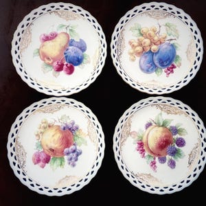 May include: Set of four white porcelain plates with a scalloped edge and gold trim. Each plate features a different hand-painted fruit design: pear, grapes, plums, and peach. Decorative tableware.
